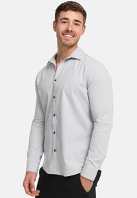 Lichtgrijze lange mouwen shirt gemaakt van stof met een subtiele textuur, met een knoopsluiting aan de voorkant en bruine knopen, met een open kraag.
