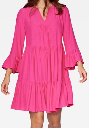 Femme portant une robe rose vif, à manches longues avec poignets évasés, longueur au genou, décolleté en V et ourlet à volants à étages.