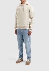 Pullover hoodie van lichtbeige katoen, met een kangoeroezak en een capuchon met trekkoord, gecombineerd met blauwe denim jeans en bruine sneakers.
