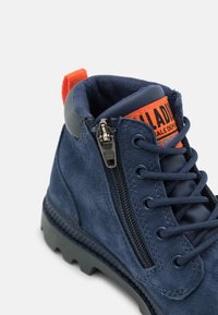 Navy modré semišové kotníkové boty se zipem na boku, kulatými tkaničkami a robustní gumovou podrážkou. Na límci se nachází oranžová značka.