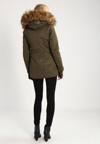Žena v olivově zelené parce s kapucí lemovanou kožešinou, černých skinny kalhotách a černých vysokých botách, stojící zády k bílé pozadí.