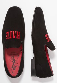 Zwarte suède loafers met rode geborduurde tekst "HATE" op de bovenkant. De binnenkant heeft een rode zool met een grafisch ontwerp. Platte hak.