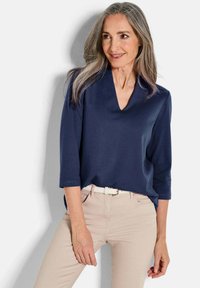 Donkerblauwe v-hals top met driekwartmouwen, een soepele textuur en een ontspannen pasvorm, gecombineerd met lichtbeige broeken en een witte riem.