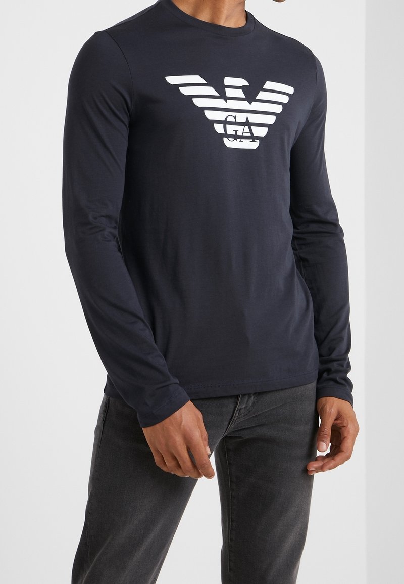 Marinblå långärmad skjorta i bomull, med en vit randig logotyp på bröstet. Enkel design, rund halsringning, mjuk textur.