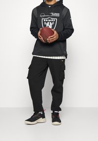 Černá mikina s kapucí s logem Las Vegas Raiders, pruhovanými detaily a klokankou; spárovaná s černými cargo kalhotami a sportovními teniskami.
