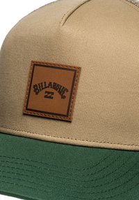 Gorra marrón y verde con un panel frontal beige, que presenta un parche rectangular de cuero grabado con "Billabong" y un logotipo.