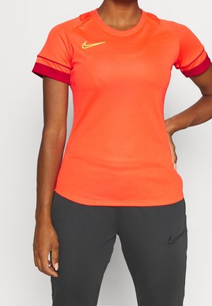 Person trägt ein knallorangafarbenes Sportshirt mit kurzen Ärmeln und gelbem Logo sowie dunkelgraue Hose mit schwarzem Logo und steht mit einer Hand in der Hüfte.