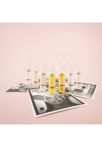 Conjunto de ocho ampollas de vidrio en varios colores (transparente, rosa, amarillo) etiquetadas con nombres de productos, colocadas sobre material impreso en blanco y negro contra un fondo rosa.