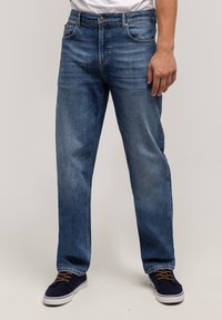 Niebieskie dżinsy z denimu o prostym kroju, z pięcioma kieszeniami, zapięciem na guziki i lekkim spraniem. Stylizowane z granatowymi butami i białym t-shirtem.