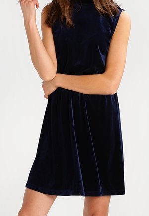 Kobieta w granatowej aksamitnej sukience bez rękawów, z jedną ręką założoną na drugą i drugą dłonią dotykającą twarzy.