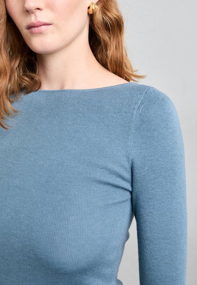 Pulover ribbed de culoare albastru deschis, cu un decolteu tip barcă și mâneci lungi, care evidențiază o textură moale și un design ajustat. Cercei aurii vizibili.