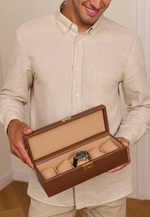 Man in beige shirt holding open brown watch case with one black wristwatch inside on cushioned slot.