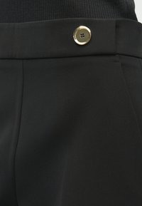 Close-up de calças pretas com um botão com detalhes dourados e detalhe de bolso, usadas com um top preto canelado.