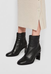 Botas de tobillo de cuero negro con punta afilada, tacón alto de bloque y cremallera lateral, combinadas con una falda de canalé beige con un detalle de botón.