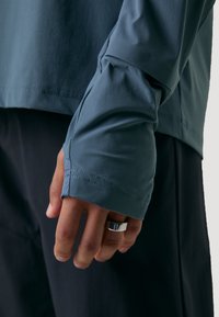 Langemouwen top in gedempt blauwe stof met een gladde textuur. Heeft strakke manchetten en minimale stiksels, gecombineerd met donkere pantalon.
