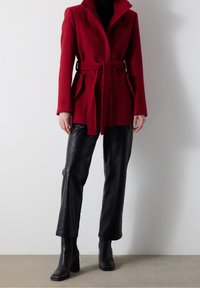 Abrigo de lana rojo con cinturón en la cintura, cuello alto y bolsillos laterales, combinado con pantalones de cuero negro acolchados y botas negras de tacón cuadrado.