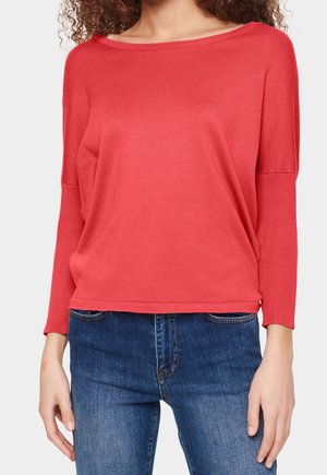 Žena v lososově růžovém tričku s dlouhým rukávem a výstřihem do lodičky a modrých džínách stojí s rukama volně podél těla.