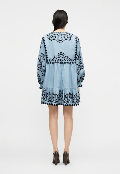 Γυναίκα που φοράει ανοιχτό μπλε φόρεμα με μαύρη κεντητή λουλουδάτη διακόσμηση, μακριά μανίκια και κυματιστά περιθώρια, σε συνδυασμό με καφέ παπούτσια με ψηλό τακούνι, γυρισμένη προς τα πίσω.