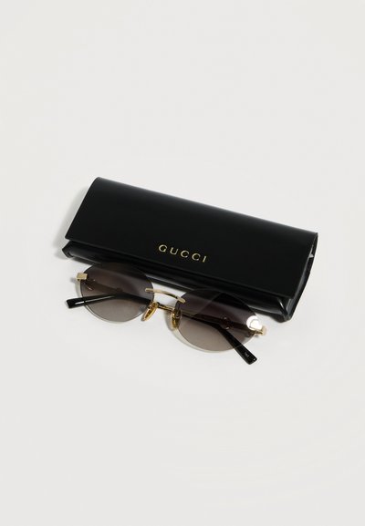Γυαλιά ηλίου με στρογγυλούς φακούς και χρυσό σκελετό δίπλα σε μαύρη θήκη Gucci με χρυσά γράμματα σε απλό ανοιχτό φόντο.