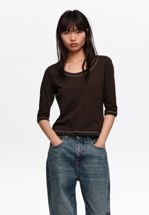 Joven con cabello lacio y negro, lleva una camisa marrón oscuro de tres cuartos de manga y jeans azules sueltos, con las manos en los bolsillos, fondo neutro.