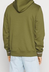 Sudadera verde oliva con capucha ajustable, puños acanalados con rayas y un corte relajado, combinada con jeans de color azul claro.
