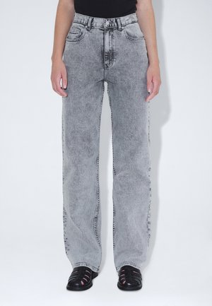 Person iført lysegrå højttaljede jeans med lige ben og sorte lukkede sandaler, stående foran en ensfarvet hvid baggrund.