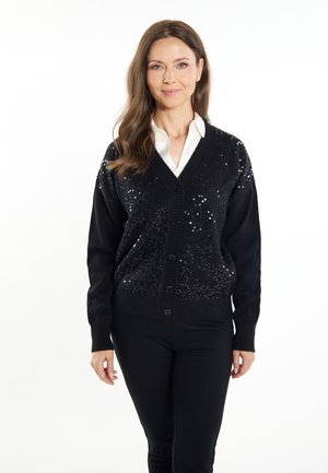 Cardigan nero con scollo a V, caratterizzato da paillettes sulla parte anteriore. Tessuto morbido, polsini e orlo a costine, e chiusura con bottoni. Indossato sopra una camicia bianca.