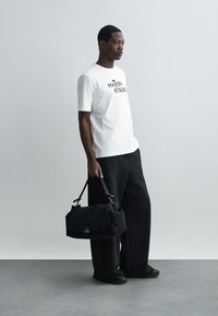 Balta kokvilnas T-kreklus ar "MAISON KITSUNÉ" logotipu, kombinēts ar melniem brīviem biksēm. Tur melnu, teksturētu auduma somu ar logotipa detaļu.