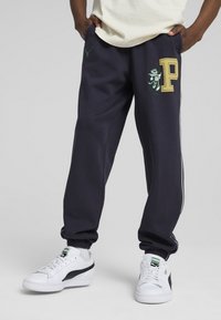 Pantaloni jogger blu navy con elastici alle caviglie e tasche, con un grande logo dorato "P" e un design di leone ricamato in verde. Abbinati a sneakers bianche.