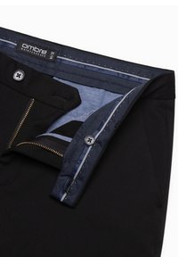 Musta housut, joissa on vetoketju, kultainen vetoketju ja tummansininen kuvioitu vuori, napin suljinto ja etiketti, jossa lukee "ombre quintessence."