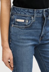 Μπλε τζιν παντελόνι με στενή εφαρμογή και διακριτική ξεθώριαση, με ετικέτα λογοτύπου Calvin Klein στην μπροστινή τσέπη και ασημένιες μεταλλικές λεπτομέρειες.