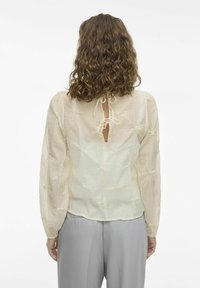 Jasno beżowa bluzka z przezroczystego materiału, z bufkami i dekoracyjnymi węzłami. Posiada związaną otwartą plecy i luźny krój.