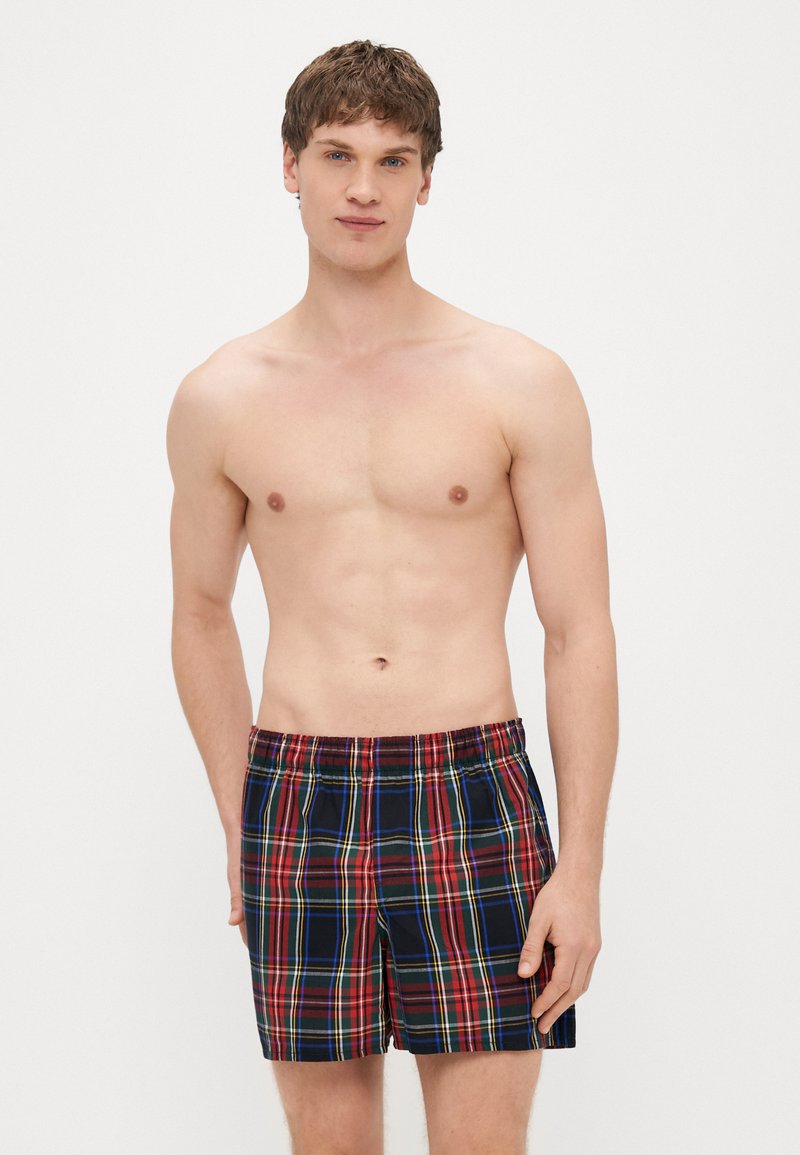 Kąpielówki w kratkę z szerokim elastycznym pasem, zdobione czerwonymi, zielonymi i żółtymi liniami na granatowym tle. Lekka tkanina, długość do kolan.