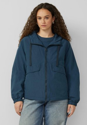 Vrouw met krullend haar, gekleed in een blauw ritssluitingvest met capuchon en lichtgewassen spijkerbroek, staand tegen een effen grijze achtergrond.