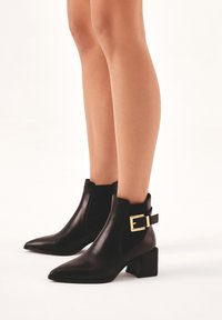 Black leather ankle boots with a pointed toe, elastic side panels, and a gold buckle accent on the ankle strap. Block heel design.