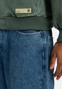 Sudadera verde con puños y dobladillo de canalé, etiqueta con la inscripción "EST. 1992". Combinada con jeans de mezclilla azules de corte clásico y detalles en los bolsillos.