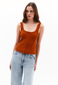 Top a canottiera a coste color ruggine con scollatura a U, chiusura frontale con bottoni e design aderente. Abbinata a jeans a gamba larga blu chiaro.