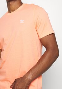 Muž v světle oranžovém tričku Adidas s bílým logem vlevo na hrudi, s uvolněnou rukou podél těla na bílém pozadí.