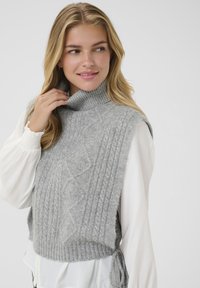 Sivi pleteni turtleneck prsluk s rebrastim teksturom i uzorkom pletenice. Bijela bluzica s nabranim rukavima vidi se ispod.