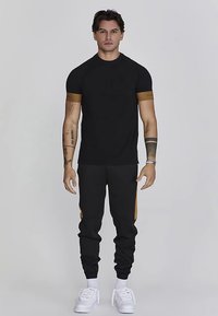 Črna majica s kratkimi rokavi z rjavimi poudarki in oprijete črne športne hlače, opremljene z bočnimi paneli, elastičnimi manšetami ter belimi supergami.