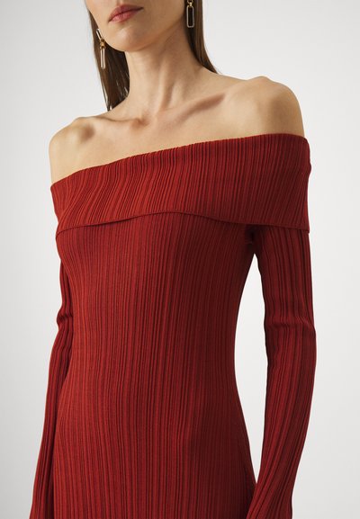Femeie îmbrăcată într-o rochie roșie, cu mâneci lungi, cu un design cu dungi și umerii lăsați, purtând cercei dreptunghiulari aurii, pe un fond simplu.
