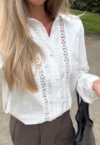 White button-up shirt featuring intricate lace detailing along the front, long puffed sleeves, and a relaxed fit, paired with brown trousers.