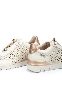 Zapatillas de cuero blanco con detalles dorados, diseño perforado, punta redondeada y suela gruesa blanca. Presenta un cierre con cordones y elementos decorativos.