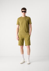 Set format dintr-un tricou din bumbac de culoare verde măslin și pantaloni scurți, prezentând mâneci scurte, guler rotund și logo brodat, asortat cu adidași albi și ochelari de soare.