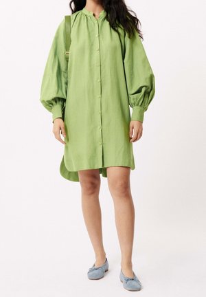 Femme portant une robe verte ample à boutons avec des manches ballon, portant un sac à bandoulière vert assorti et des ballerines bleu clair.