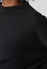 Tissu noir texturé avec une surface légèrement froissée ; présente un col montant et un drapé décontracté, mettant en valeur un motif subtil et irrégulier.