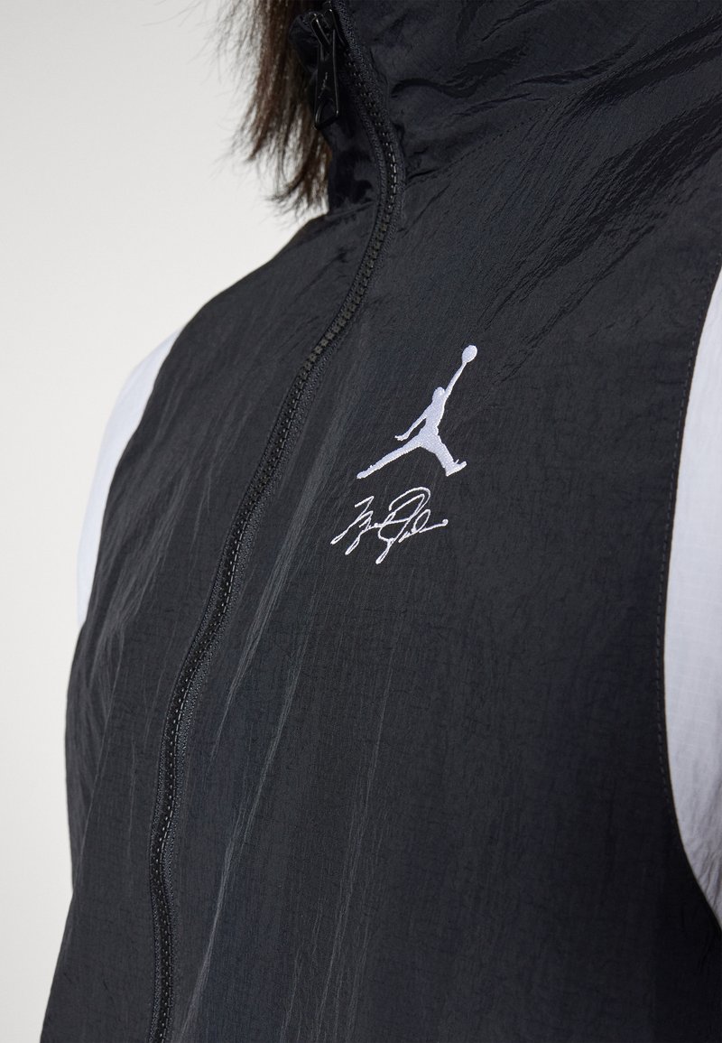 Veste légère noire avec une fermeture éclair à l'avant, comportant un logo Jumpman blanc et une signature sur la poitrine gauche, avec des accents blans contrastants sur les épaules.