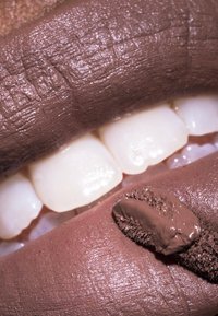 Produit à lèvres marron mat sur une surface brune texturée avec des dents blanches partiellement visibles. L'applicateur a un design élégant et une formule épaisse.