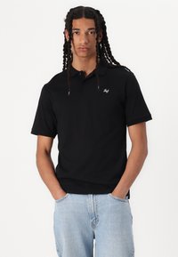 Svart kortermet polo skjorte med krage, med en subtil brodert logo på brystet og en glatt bomullstekstur. Brukt med lyseblå jeans.