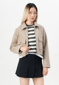 Beige zip-up jacket with a collar, two front pockets, worn over a striped green and white shirt, paired with a black pleated skirt.